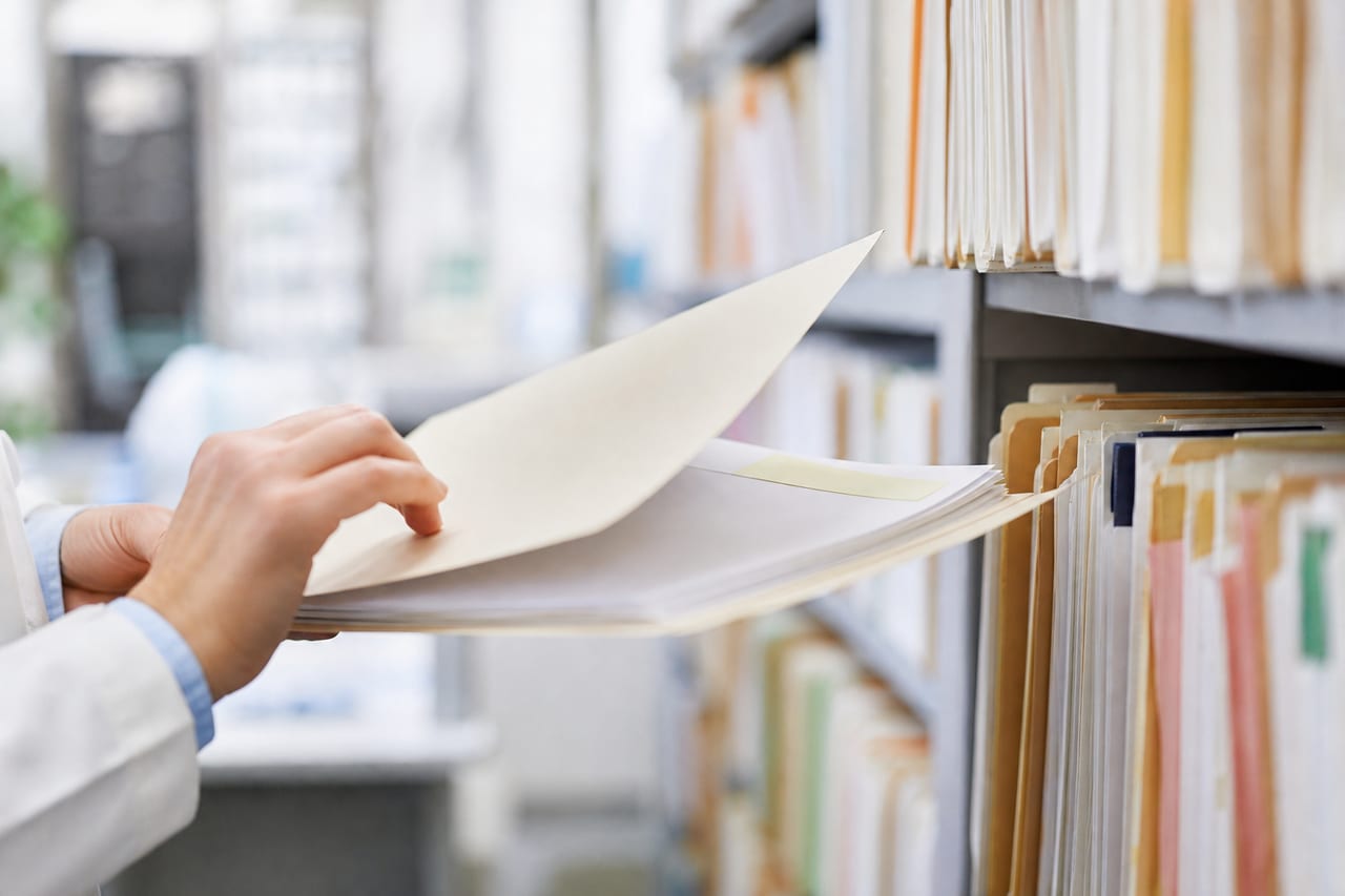 Bureau des archives médicales d’un hôpital, dossiers papier et classeurs sur une étagère, professionnel de santé consultant un document, lumière naturelle